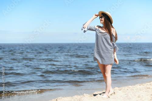 Happy young brunette woman standing on the ocean beach while smiling, and wearing fashion hat, sunglasses. The enjoying vacation concept.