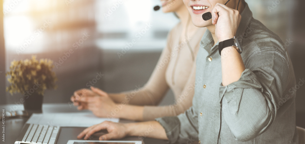 Fototapeta premium Two smiling people in headsets are talking to the clients, while sitting at the desk in sunny modern office.
