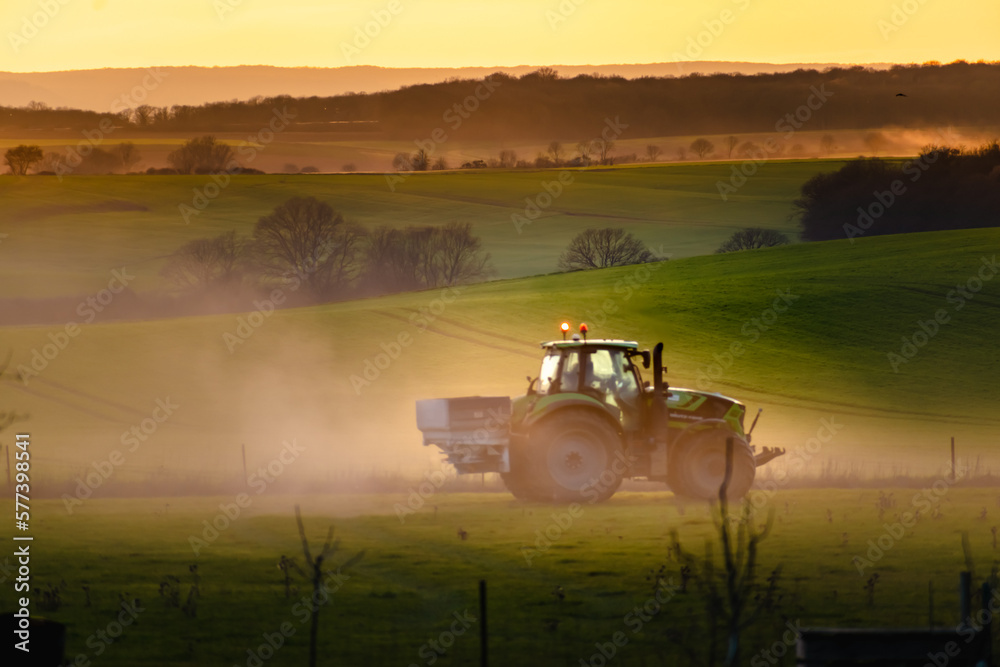 Fototapeta premium Tractor in farmland working on soil field with sunset, big equipment, countryside scene