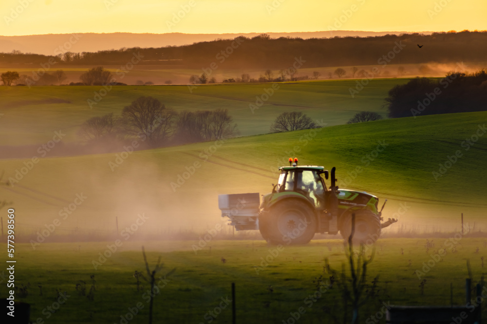 Fototapeta premium Tractor in farmland working on soil field with sunset, big equipment, countryside scene