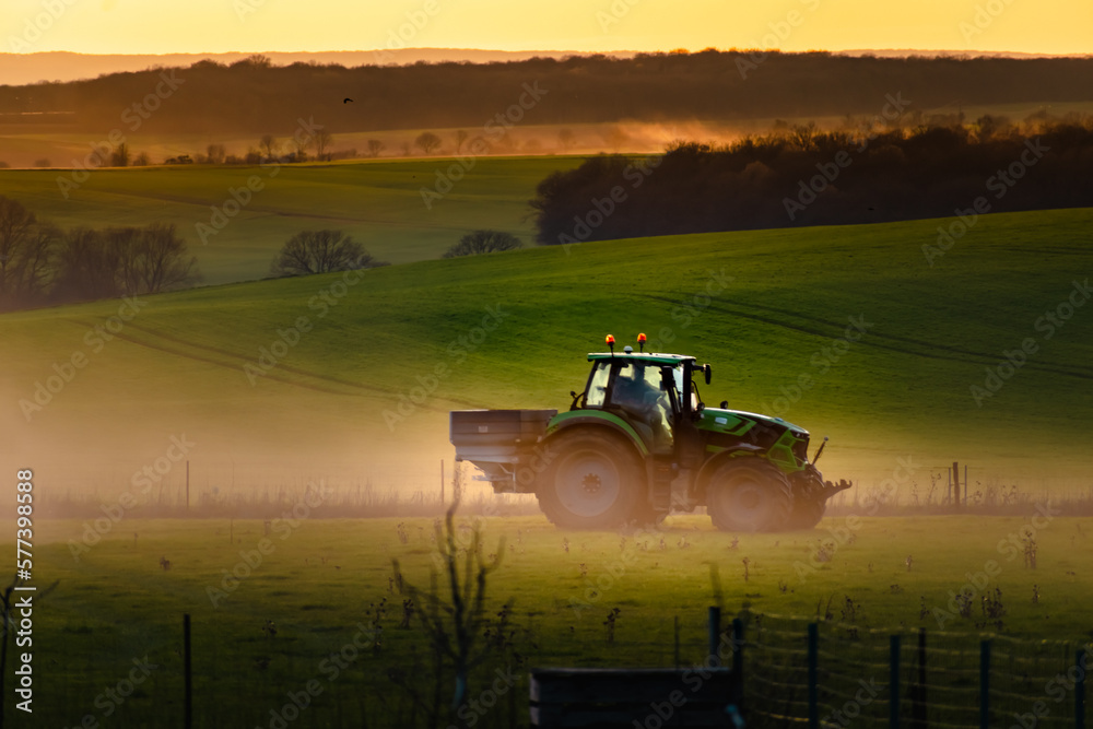 Fototapeta premium Tractor in farmland working on soil field with sunset, big equipment, countryside scene