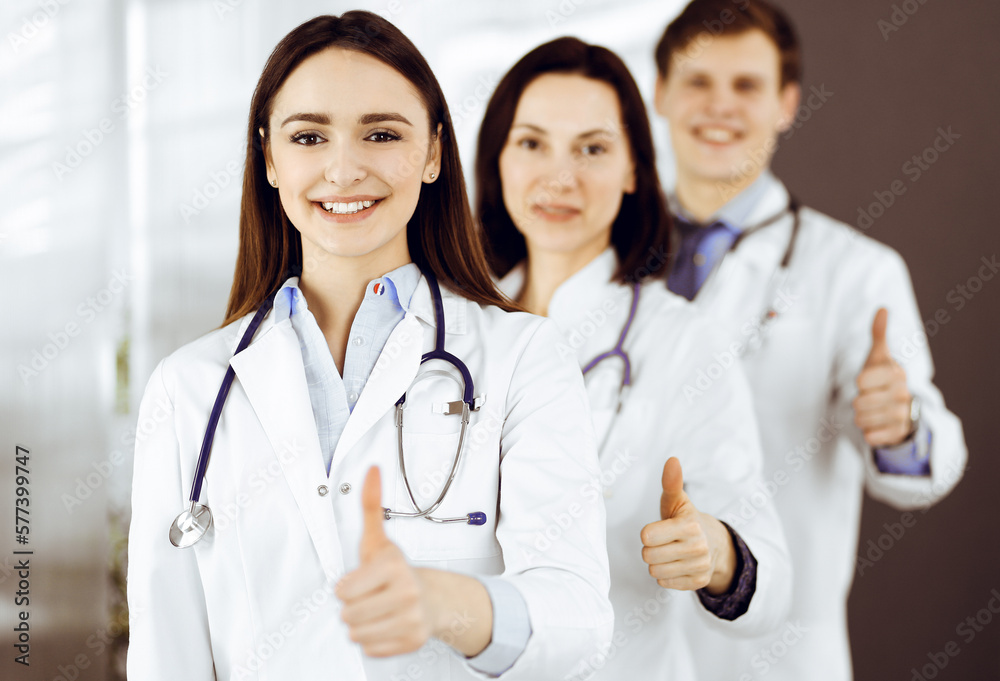 Group of young cheerful doctors is standing as a team with thumbs up in a hospital office and is ready to help patients. Medical help, insurance in health care Group of young cheerful doctors is standing as a team with thumbs up in a hospital office and is ready to help patients. Medical help, insurance in health care