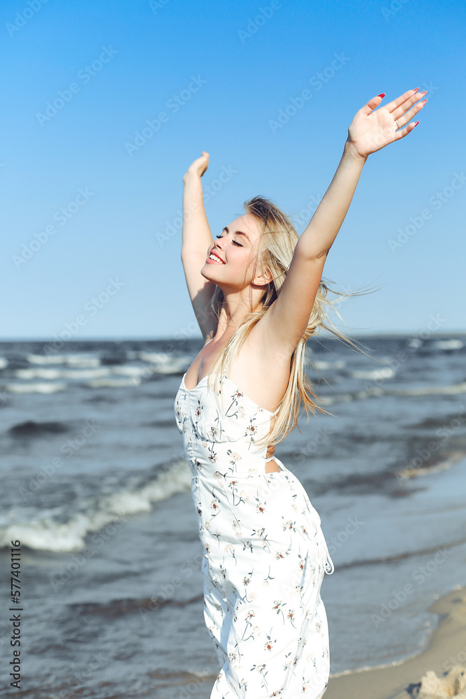 Happy blonde beautiful woman on the ocean beach standing in a white summer dress, raising hands. Happy blonde beautiful woman on the ocean beach standing in a white summer dress, raising hands.