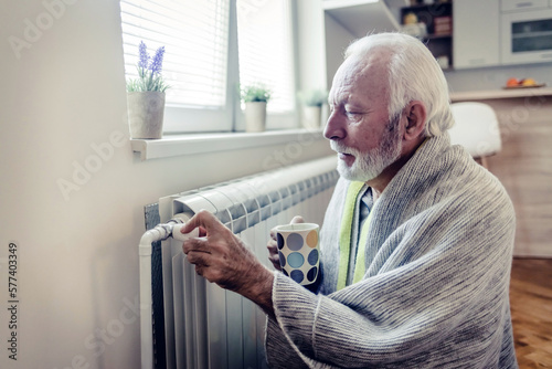 Mature man wrapped in a blanket adjusting the radiator thermostat. Sebior male adjusting the temperature on the radiator at home.