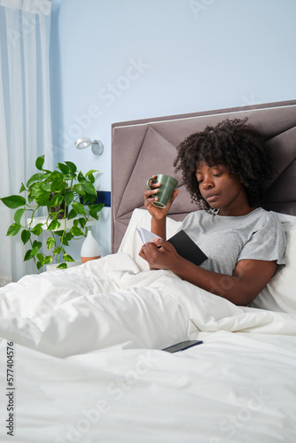 African young woman reading a book and drinking coffee in bed in the morning.