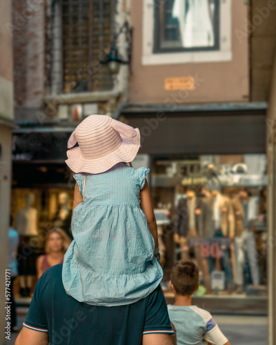 A little girl in a summer dress and a straw hat on her father's shoulders on the street of an old European city, a boy walks ahead against a blurred background of shop windows. Family travel
