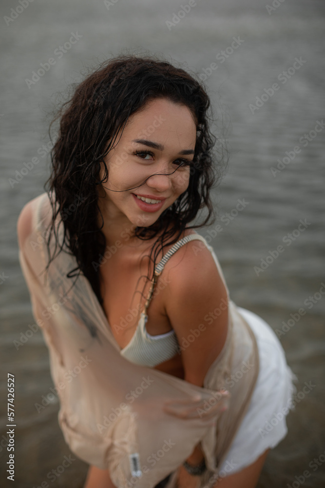 Charming happy young woman with wet hair after bathing in white bikini poses standing in water and looking at camera. 