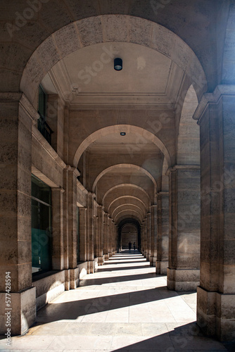 Paris ancient stone building colonnade with massive columns, arches, stucco fretwork, stone pavement, bright sun glare between the columns and two human figures at the end