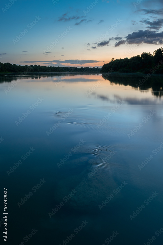 Underwater boulders in river at dusk, landscape of Sava river with forest silhouette on shore and clouds reflecting in water