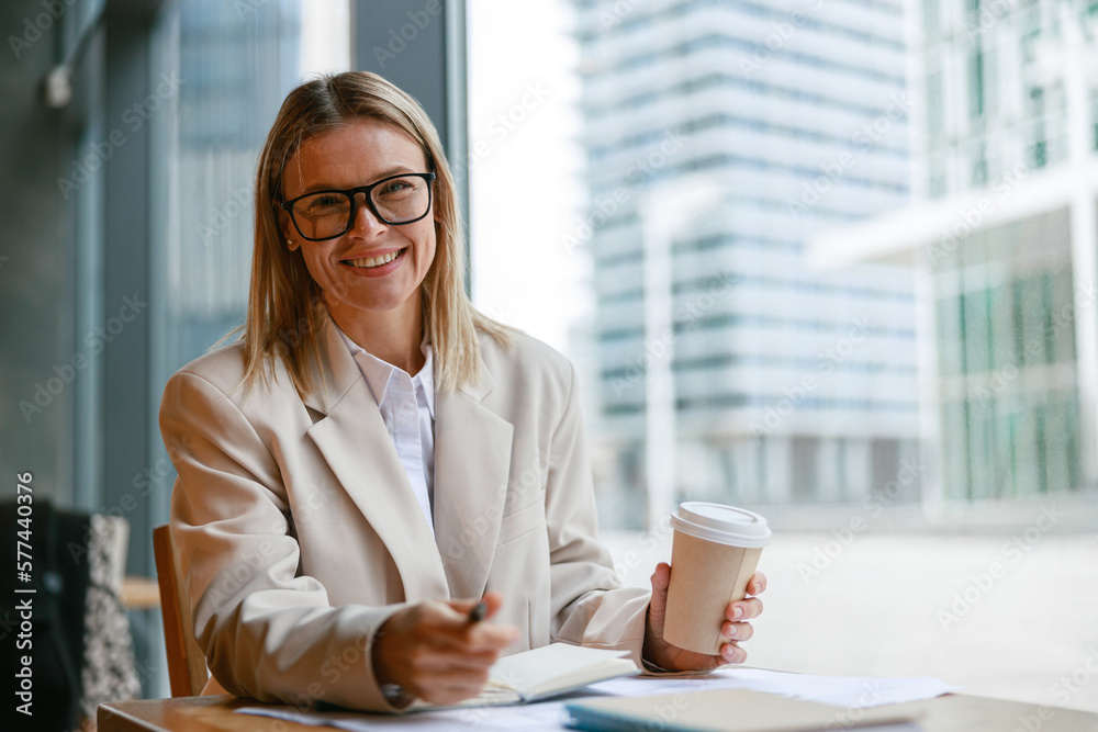 Smiling businesswoman in glasses drinking coffee and making notes while working in cafe Smiling businesswoman in glasses drinking coffee and making notes while working in cafe