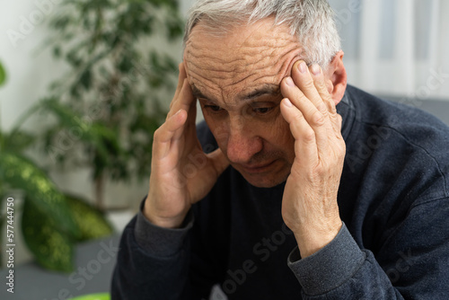 Senior man having headache and touching his head while suffering from a migraine in the living room, Mature man presses a hand to head suffers from unbearable pain.