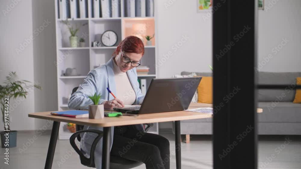 telecommuting, female manager works at computer writes data in notebook while sitting at table indoors telecommuting, female manager works at computer writes data in notebook while sitting at table indoors