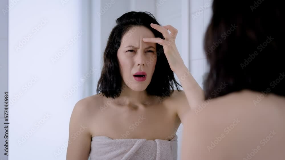 Upset middle aged woman looking at her wrinkles in bathroom mirror. Frustrated