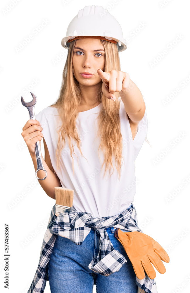 Beautiful caucasian woman with blonde hair wearing hardhat and builder clothes pointing with finger to the camera and to you, confident gesture looking serious