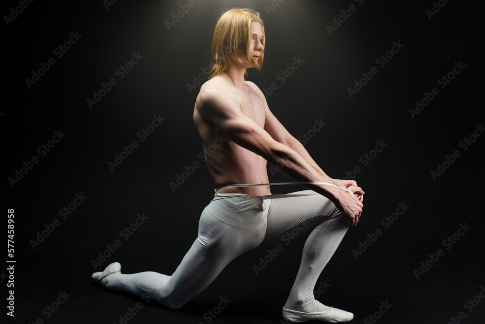 Young athletic professional ballet dancer with a bare torso and white dance tights is in perfect shape, performing and posing over a black background.