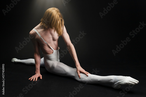 Young athletic professional ballet dancer with a bare torso and white dance tights is in perfect shape, performing and doing splits over a black background.