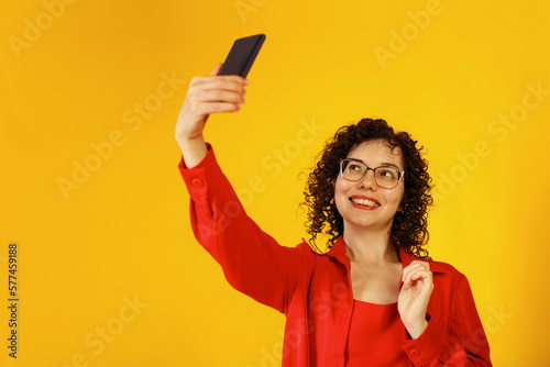 Cute curly girl with glasses taking a selfie. A young woman takes a picture of herself on the phone. Studio female portrait on a bright yellow background.
