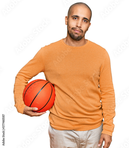 Hispanic adult man holding basketball ball thinking attitude and sober expression looking self confident