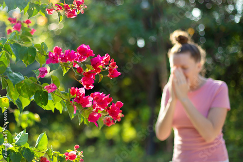 Portrait of beautiful young allergic woman is suffering from pollen allergy or cold on natural flower flowering tree background at spring or sunny summer day sneezes, blowing her runny nose rubs eyes