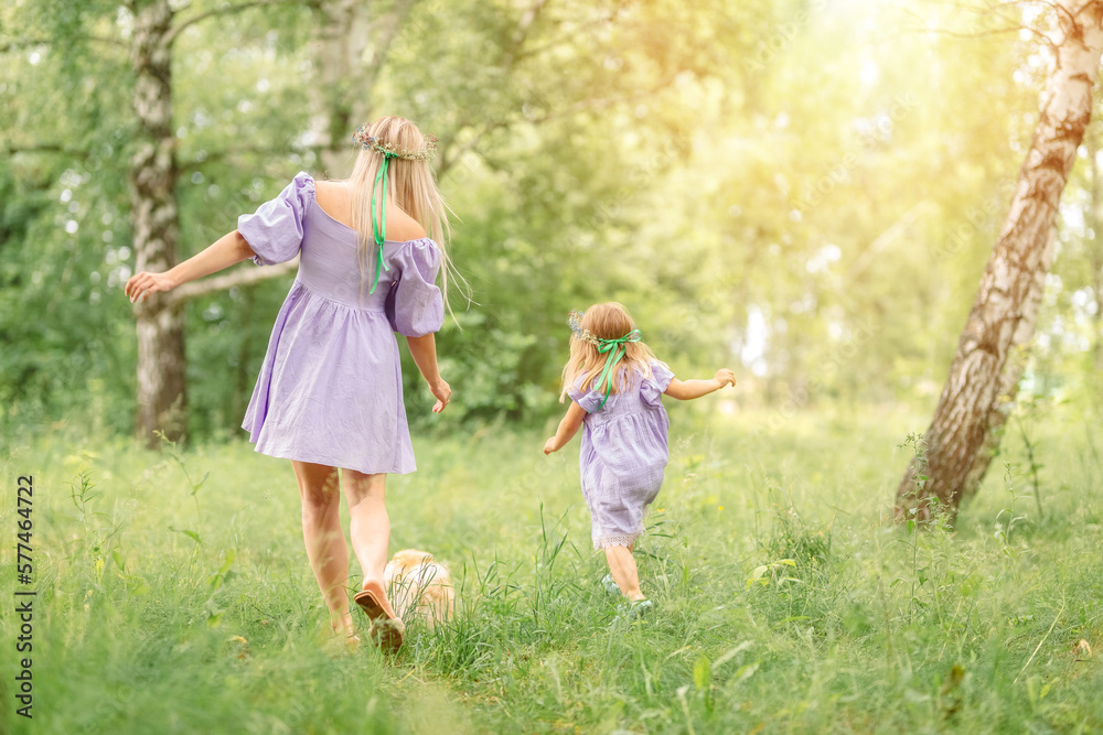 Fototapeta premium mother and daughter in short lilac dresses run along a summer meadow with green grass and trees. A small red dog runs in front of them.