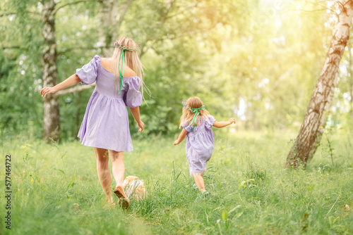 mother and daughter in short lilac dresses run along a summer meadow with green grass and trees. A small red dog runs in front of them.