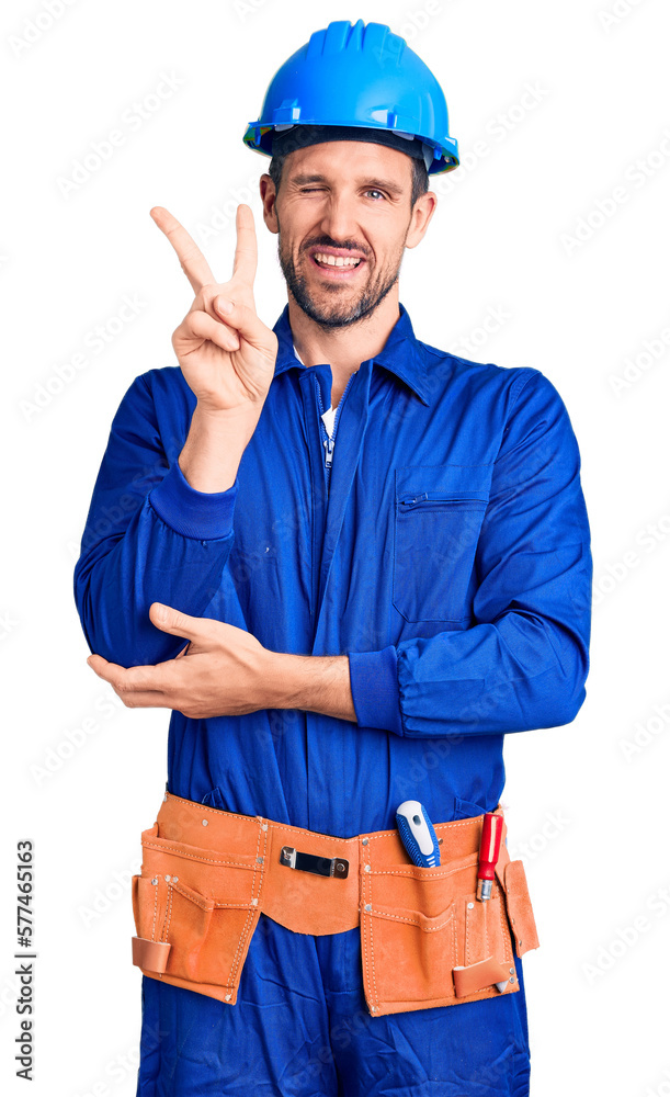Young handsome man wearing worker uniform and hardhat smiling with happy face winking at the camera doing victory sign with fingers. number two. Young handsome man wearing worker uniform and hardhat smiling with happy face winking at the camera doing victory sign with fingers. number two.