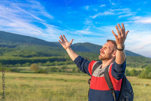 Young male traveler with a backpack raises his hands up in the background of a mountain landscape.