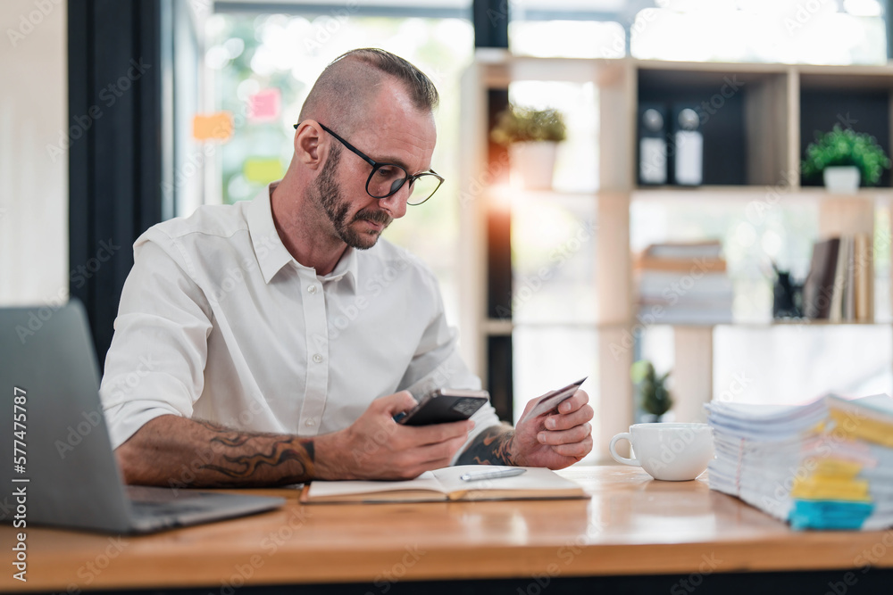 A man reading information from the back of the credit card and typing it on the smartphone to do an online purchase at home. A guy doing an online payment on a cellphone at table in office A man reading information from the back of the credit card and typing it on the smartphone to do an online purchase at home. A guy doing an online payment on a cellphone at table in office
