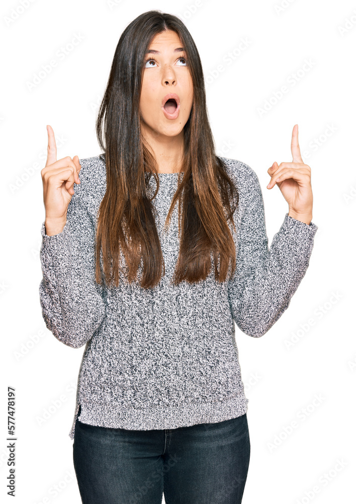 Young brunette woman wearing casual winter sweater amazed and surprised looking up and pointing with fingers and raised arms. Young brunette woman wearing casual winter sweater amazed and surprised looking up and pointing with fingers and raised arms.