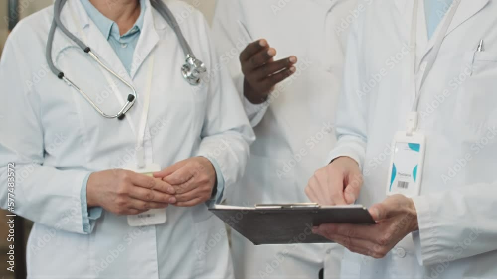 Midsection of three doctors in sterile white lab coats gesticulating while walking along hospital corridor discussing patient medical card Midsection of three doctors in sterile white lab coats gesticulating while walking along hospital corridor discussing patient medical card