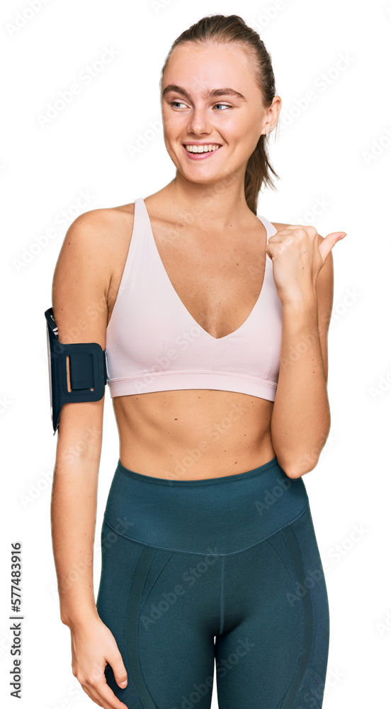 Beautiful young blonde woman wearing sportswear and arm band smiling with happy face looking and pointing to the side with thumb up. Beautiful young blonde woman wearing sportswear and arm band smiling with happy face looking and pointing to the side with thumb up.