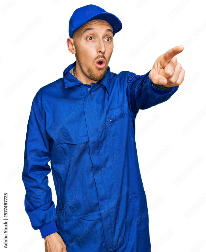 Bald man with beard wearing builder jumpsuit uniform pointing with finger surprised ahead, open mouth amazed expression, something on the front