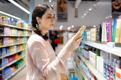 Portrait of Caucasian woman choosing cosmetic cream in grocery store. Shelves with food in background. Concept of shopping and consumerism