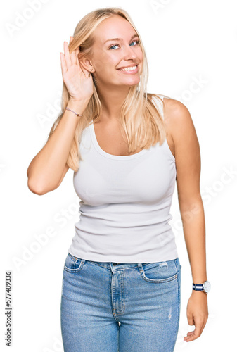 Young blonde girl wearing casual style with sleeveless shirt smiling with hand over ear listening an hearing to rumor or gossip. deafness concept.