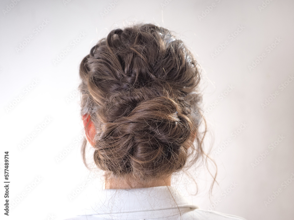  A beautiful girl gathered her wavy hair in a low bundle. Modern festive hairstyle for long brown hair. A woman braided a bunch or bun on her head on a white background. Back and side view close up