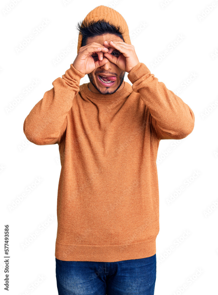 Handsome latin american young man wearing winter sweater and wool hat doing ok gesture like binoculars sticking tongue out, eyes looking through fingers. crazy expression. Handsome latin american young man wearing winter sweater and wool hat doing ok gesture like binoculars sticking tongue out, eyes looking through fingers. crazy expression.