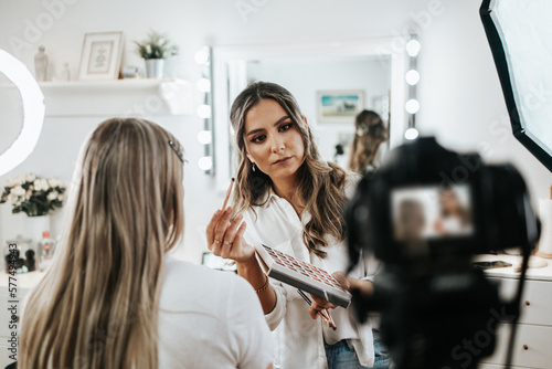 Happy young woman streaming a beauty makeup vlog from home or workshop. Beautiful online content creator cosmetician applying makeup and explaining some work tools. Vlogging and online channel work. photo