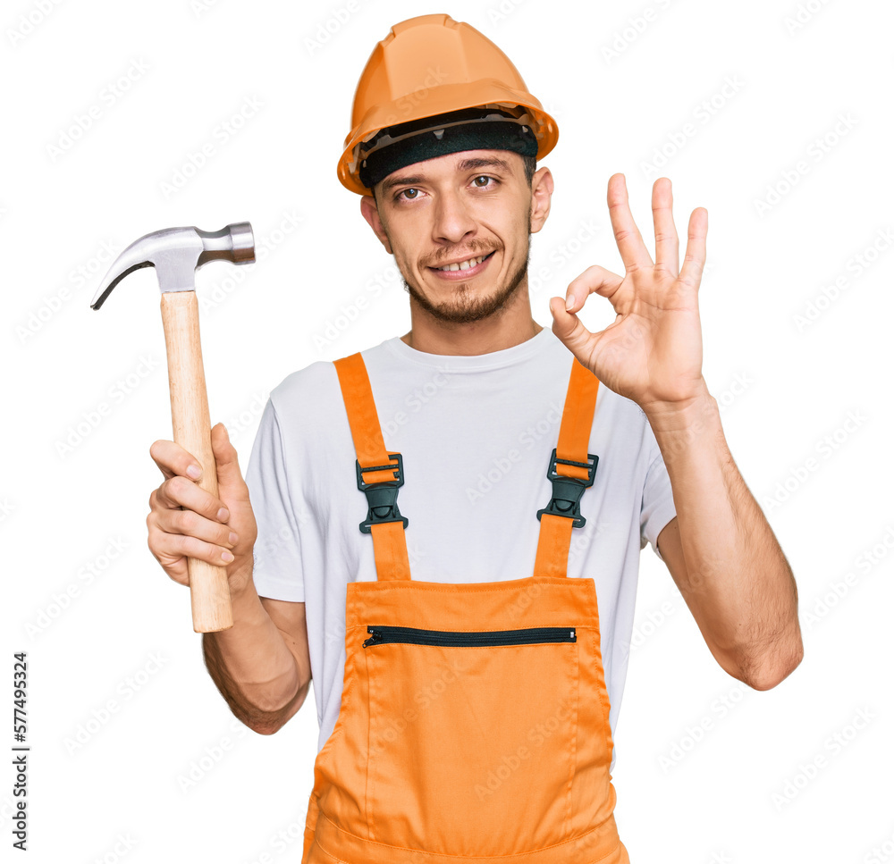 Hispanic young man wearing hardhat holding hammer doing ok sign with fingers, smiling friendly gesturing excellent symbol Hispanic young man wearing hardhat holding hammer doing ok sign with fingers, smiling friendly gesturing excellent symbol
