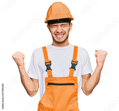 Hispanic young man wearing handyman uniform and safety hardhat very happy and excited doing winner gesture with arms raised, smiling and screaming for success. celebration concept.