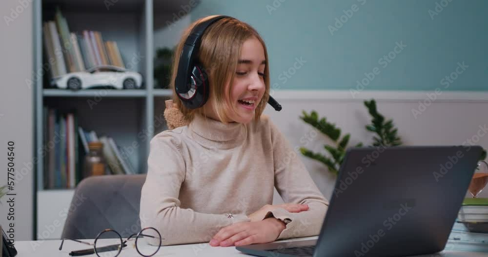 Beautiful little Caucasian girl in headphones wave hand greeting with tutor, study online lesson with teacher using laptop. Homeschooling concept