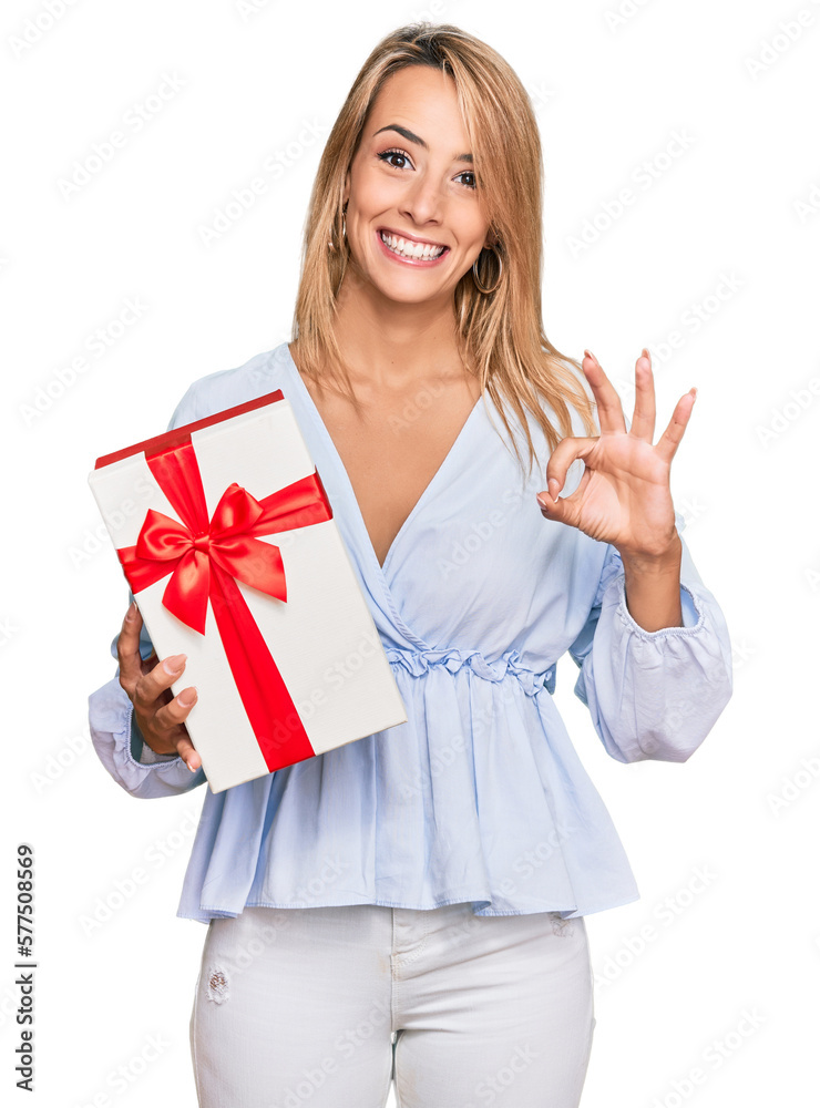 Beautiful blonde woman holding gift doing ok sign with fingers, smiling friendly gesturing excellent symbol Beautiful blonde woman holding gift doing ok sign with fingers, smiling friendly gesturing excellent symbol