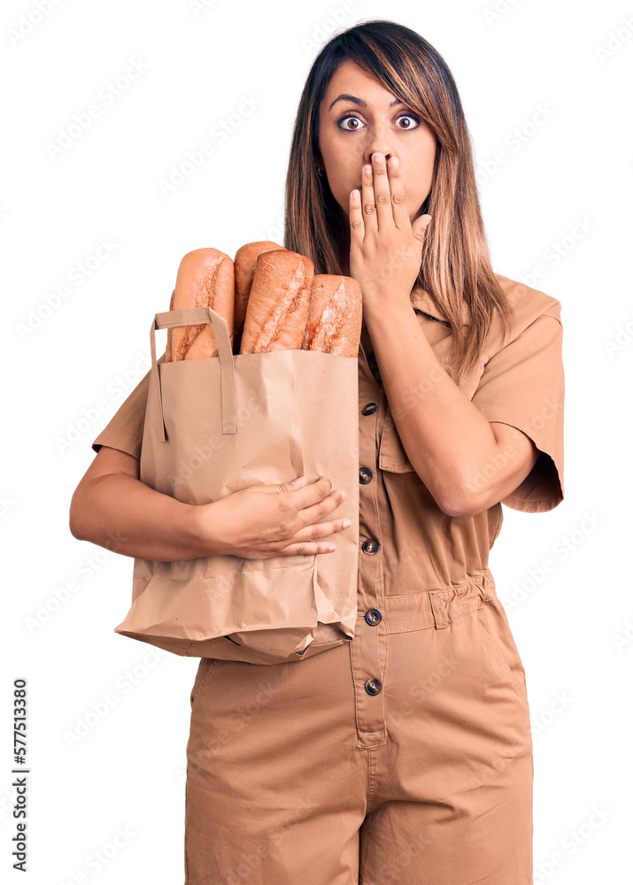 Young beautiful woman holding paper bag with bread covering mouth with hand, shocked and afraid for mistake. surprised expression Young beautiful woman holding paper bag with bread covering mouth with hand, shocked and afraid for mistake. surprised expression