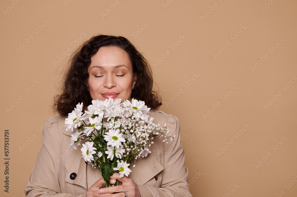 Close-up portrait of a beautiful elegant stylish Latin American woman, dressed in beige stylish coat, posing with her eyes closed and a bouquet of white spring chamomile flowers on isolated background Close-up portrait of a beautiful elegant stylish Latin American woman, dressed in beige stylish coat, posing with her eyes closed and a bouquet of white spring chamomile flowers on isolated background
