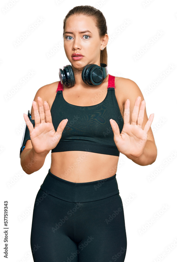 Beautiful blonde woman wearing gym clothes and using headphones moving away hands palms showing refusal and denial with afraid and disgusting expression. stop and forbidden. Beautiful blonde woman wearing gym clothes and using headphones moving away hands palms showing refusal and denial with afraid and disgusting expression. stop and forbidden.