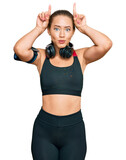 Beautiful blonde woman wearing gym clothes and using headphones doing funny gesture with finger over head as bull horns