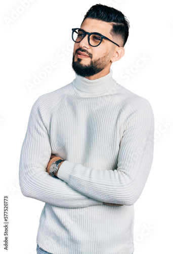 Young arab man with beard wearing elegant turtleneck sweater and glasses looking to the side with arms crossed convinced and confident