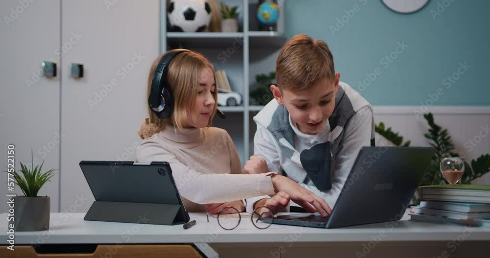 Small cute girl with headphones using internet games on digital tablet while kid boy playing game on laptop in living room at home, family concept
