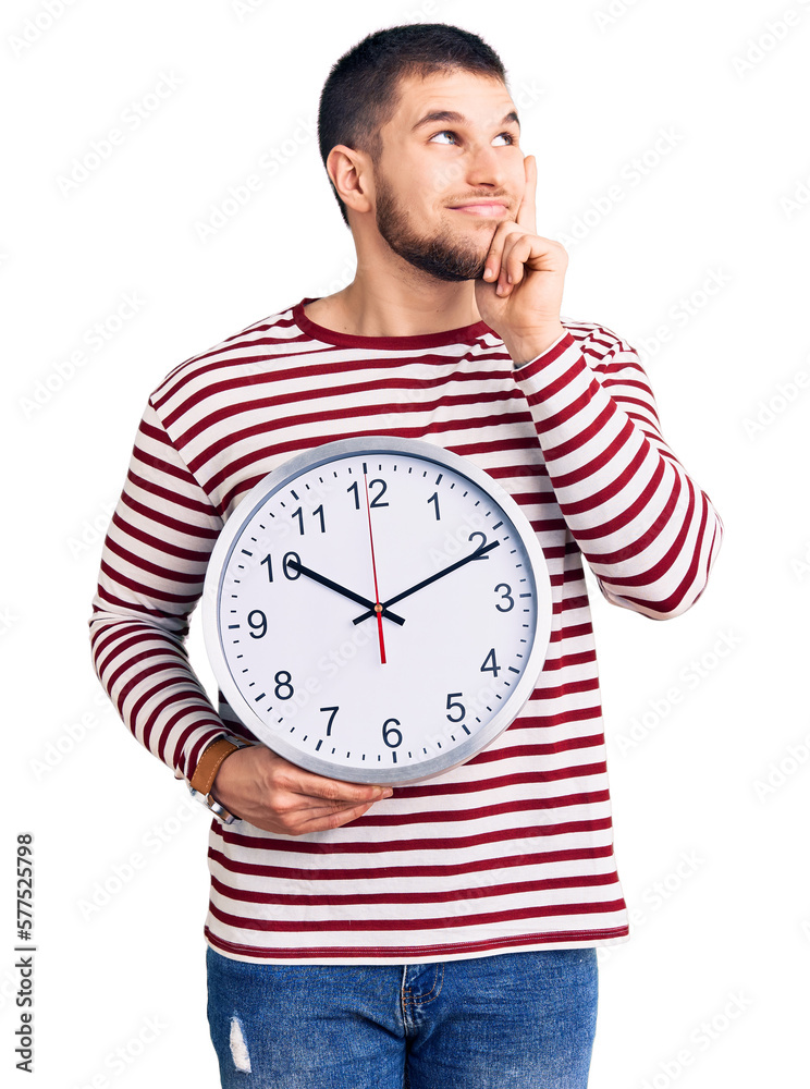 Young handsome man holding big clock serious face thinking about question with hand on chin, thoughtful about confusing idea Young handsome man holding big clock serious face thinking about question with hand on chin, thoughtful about confusing idea