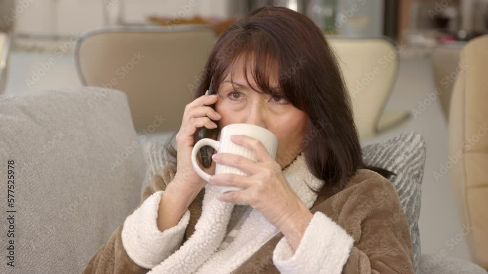 Closeup smiling attractive asian woman wearing brown white fleece collar bathrobe talking on mobile phone enjoying drinking coffee sitting, lounging on comfortable sofa in leisure home living room. Closeup smiling attractive asian woman wearing brown white fleece collar bathrobe talking on mobile phone enjoying drinking coffee sitting, lounging on comfortable sofa in leisure home living room.