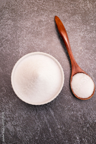 Organic sweetener Erythritol, produced by fermentation from corn, called dextrose in ceramic bowl, wooden spoon on granite background, table. Sugar substitute. Vertical plane, top view